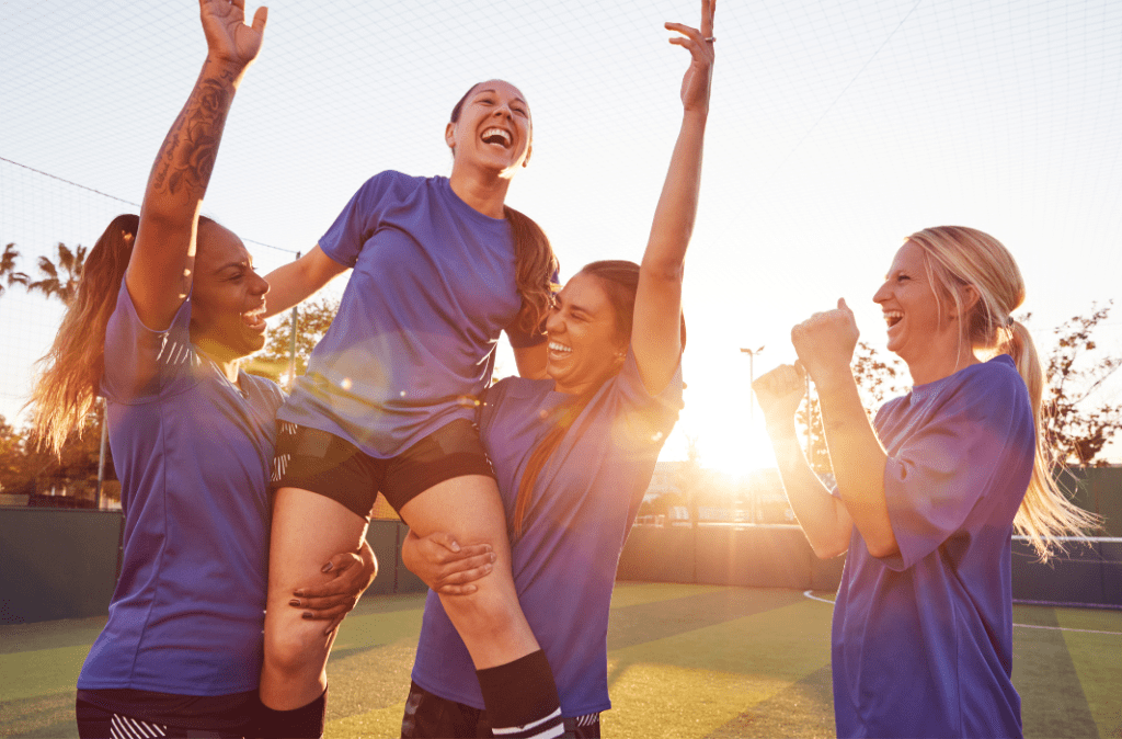 A group of four women soccer players. Two of them are left their teammate in the air as they all cheer, and the fourth woman cheers next to them. 